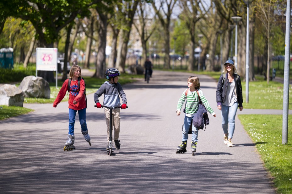 Kinderen op rolschaatsen en een step in een park samen met hun moeder
