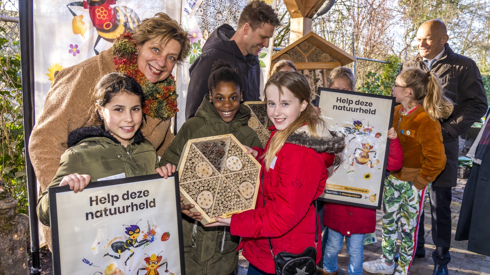 Een groep kinderen houdt posters vast waarop staat 'Help deze natuurheld' (de bij). Andere kinderen laten een bijenhotel zien. 