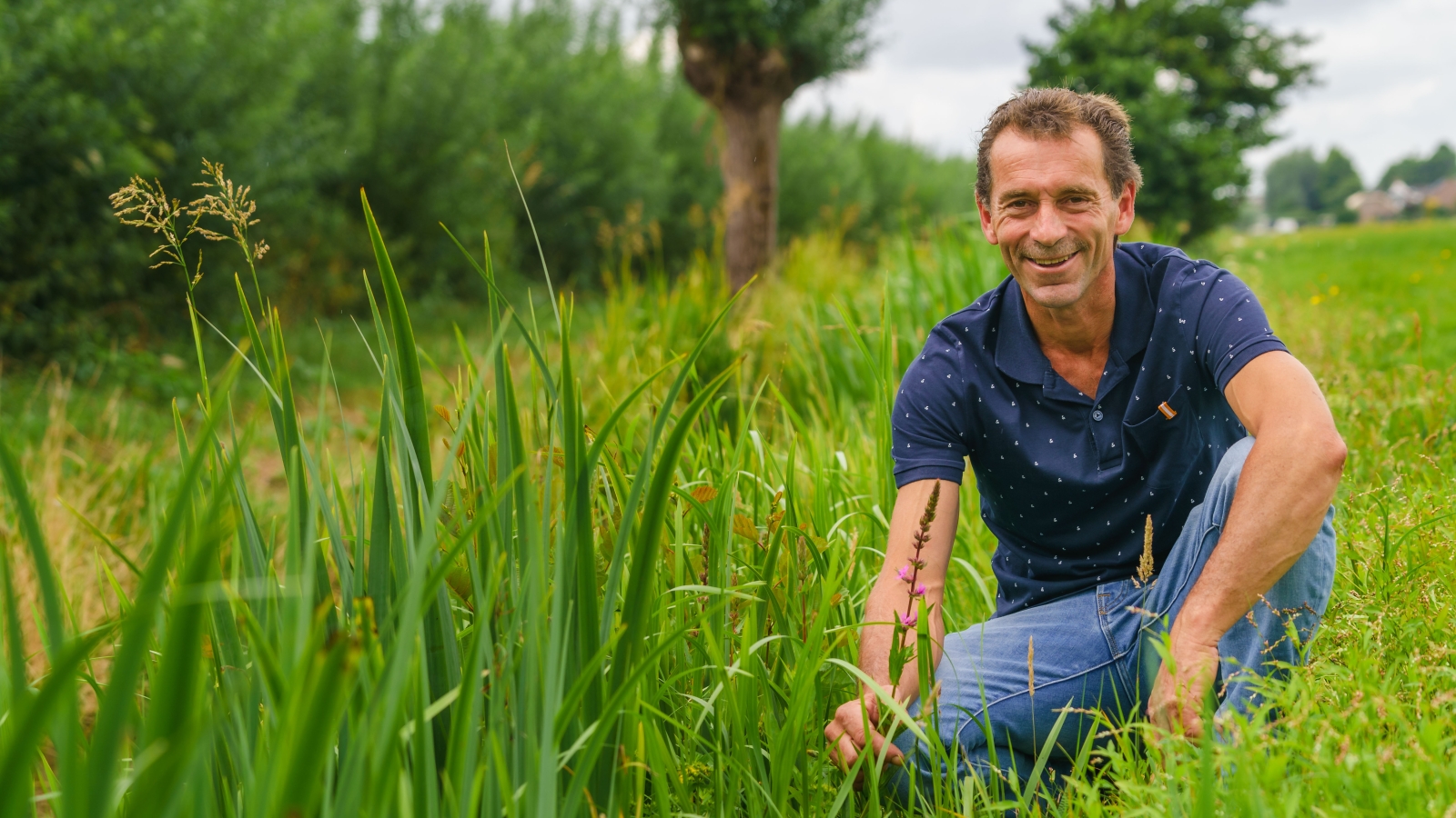 Boer net blauw shirt zit gehurkt in kruidenrijk grasland en kijkt lachend de camera in