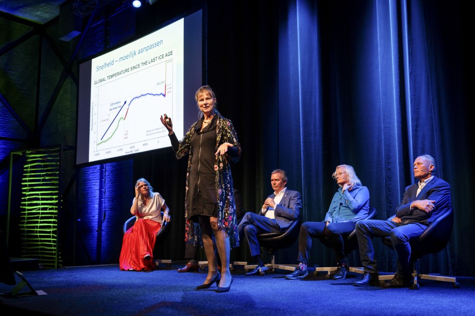 Marjan Minnesma staat op het podium met op de achergrond een scherm en vier panelleden die naar haar luisteren