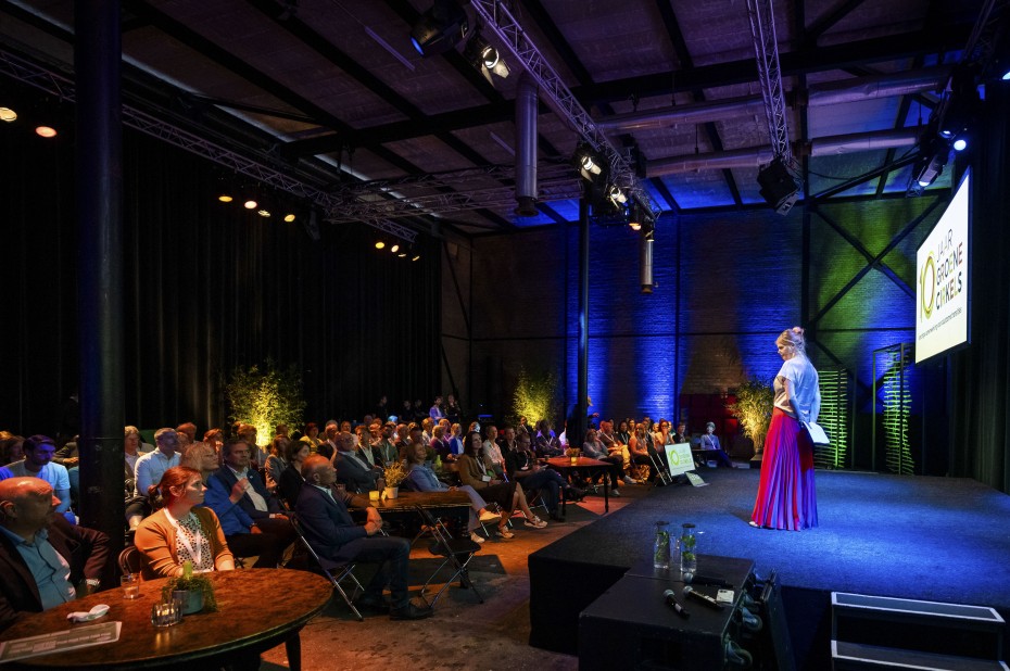 De dagvoorzitter staat met een rode rok aan op het podium en spreekt een zaal vol zittende mensen toe