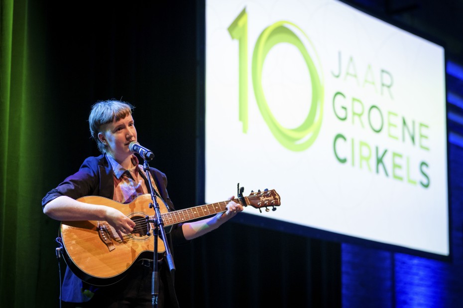 Kunstenaar Mart Nooijen zingt met een gitaar in hun hand over transitie; op de achtergrond een scherm waarop het jubileumlogo van 10 jaar Groene Cirkels te zien is