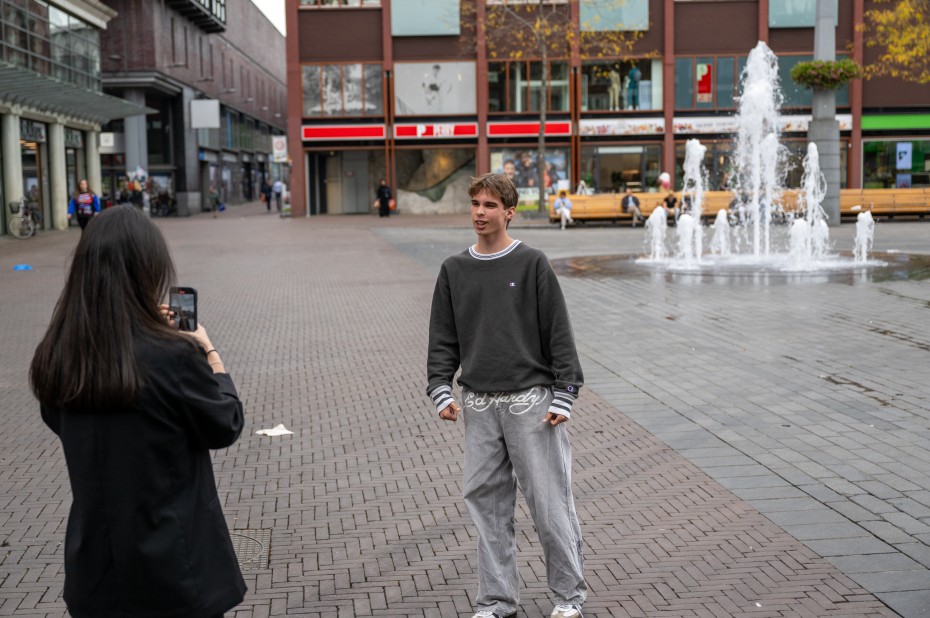 Twee jongeren nemen met hun telefoon een filmpje op op het Dordtse Statenplein. Ze staan op het plein met op de achtergrond winkels en een fontein.