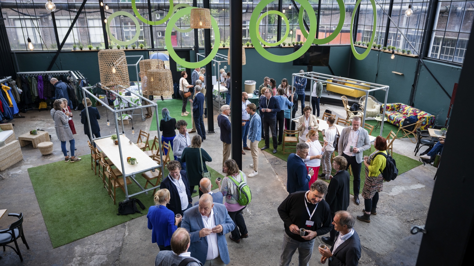 Overzichtsfoto aanwezigen jubileumevent, mensen staan te praten in een grote hal en er hangen groene cirkels aan het plafond