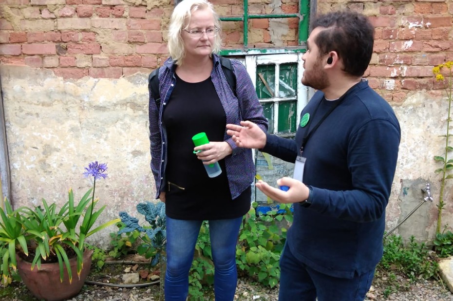 Marian Stuiver heeft een waterflesje in hun hand en staat met iemand te praten voor de gevel van een vervallen huisje