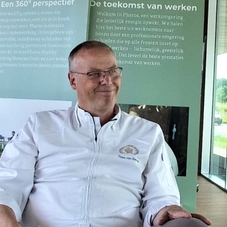Theo van Rensch zit lachend achter een bureau met op de achtergrond borden waarop staat 'De toekomst van werken'