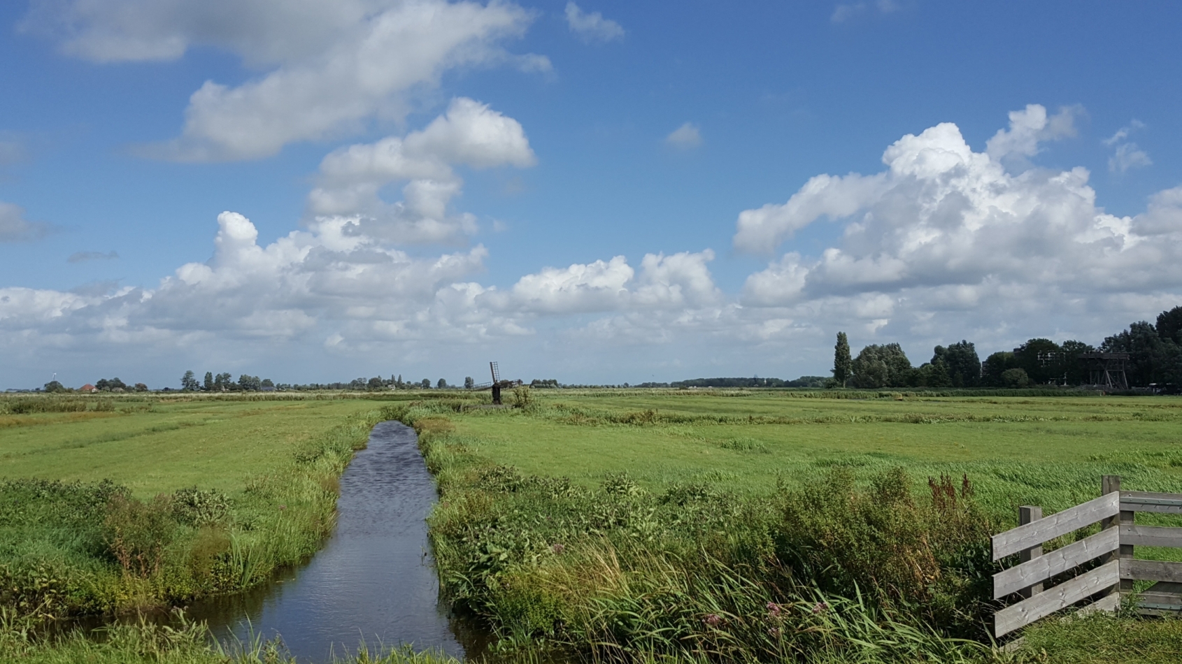 Een slootje loopt door een weiland met een hekje, de lucht is blauw met enkele wolken