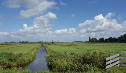 Een slootje loopt door een weiland met een hekje, de lucht is blauw met enkele wolken