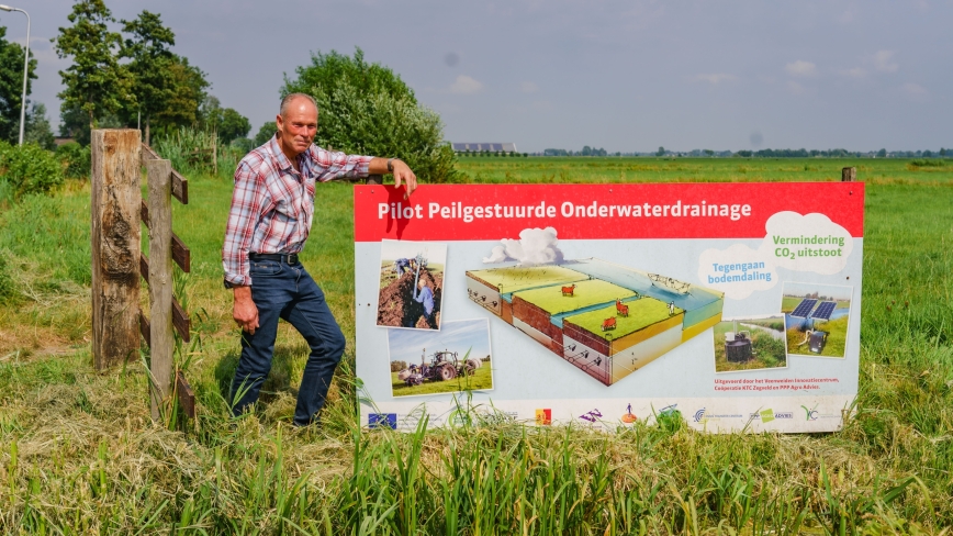 Boer staat bij een projectbord in het veld waarop staat 'Pilot peilgestuurde onderwaterdrainage'