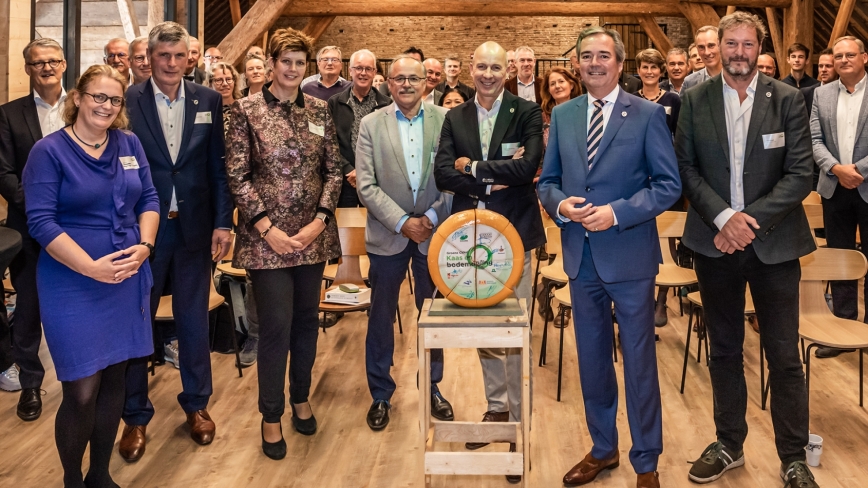 Een groep mensen in zakelijke kleding poseert naast een ronde kaas op een tafeltje