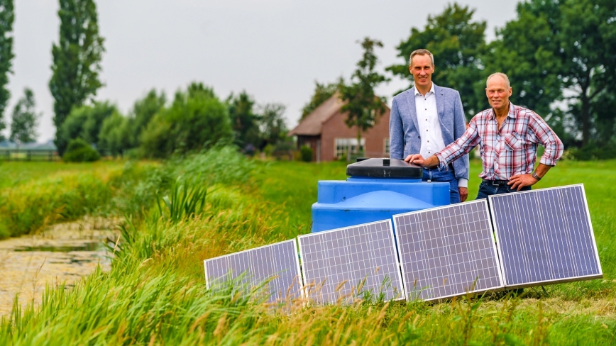 Cees van den Bos staat met veenweideboer Ad van Rees in een weiland naast zonnepanelen en een grote blauwe ton
