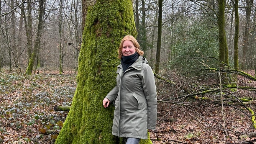 Annemarie Lammers staat met een grijze jas aan voor een boom in een bos met veel herfstbladeren op de grond